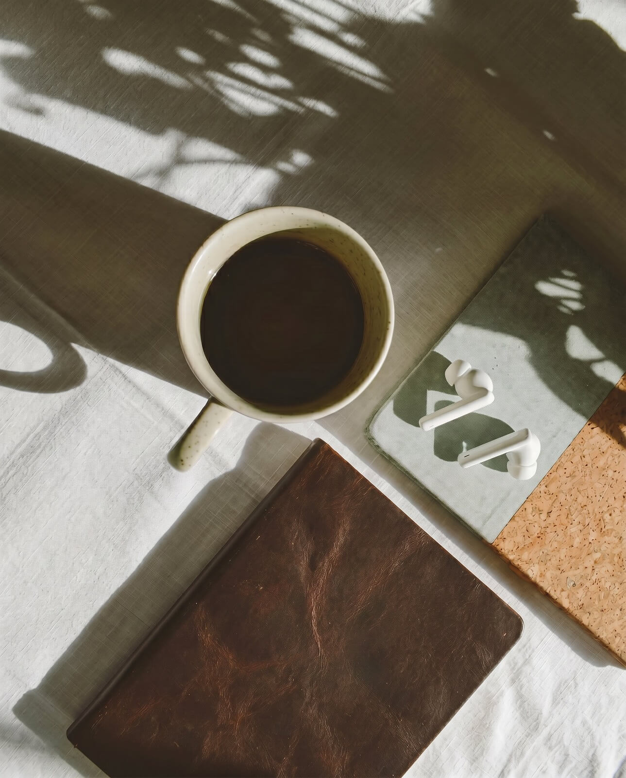 A coffee cup, leather notebook, and earbuds on rumpled linen in dappled morning light.